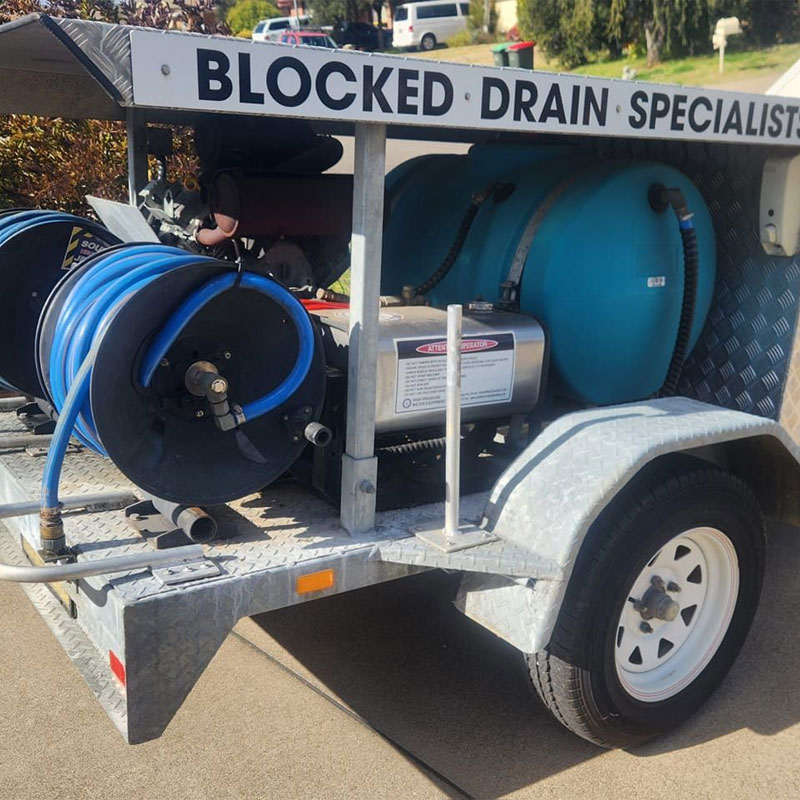 Drain jetter trailer with blue hose reel and water tank under a Blocked Drain Specialists sign